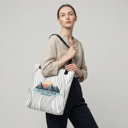 Model holding a tote with black handles featuring the “BE STILL AND KNOW — Psalm 46:10” mountain graphic on a wavy line background.