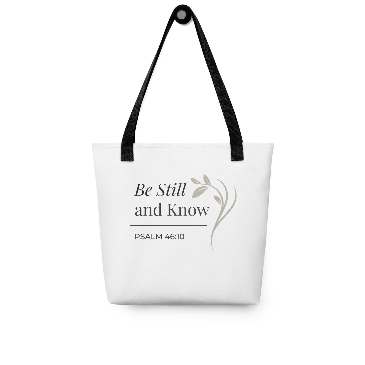 White tote bag hanging on a hook with black handles featuring “Be still and know,” “Psalm 46:10,” and a floral line design in dark gray.