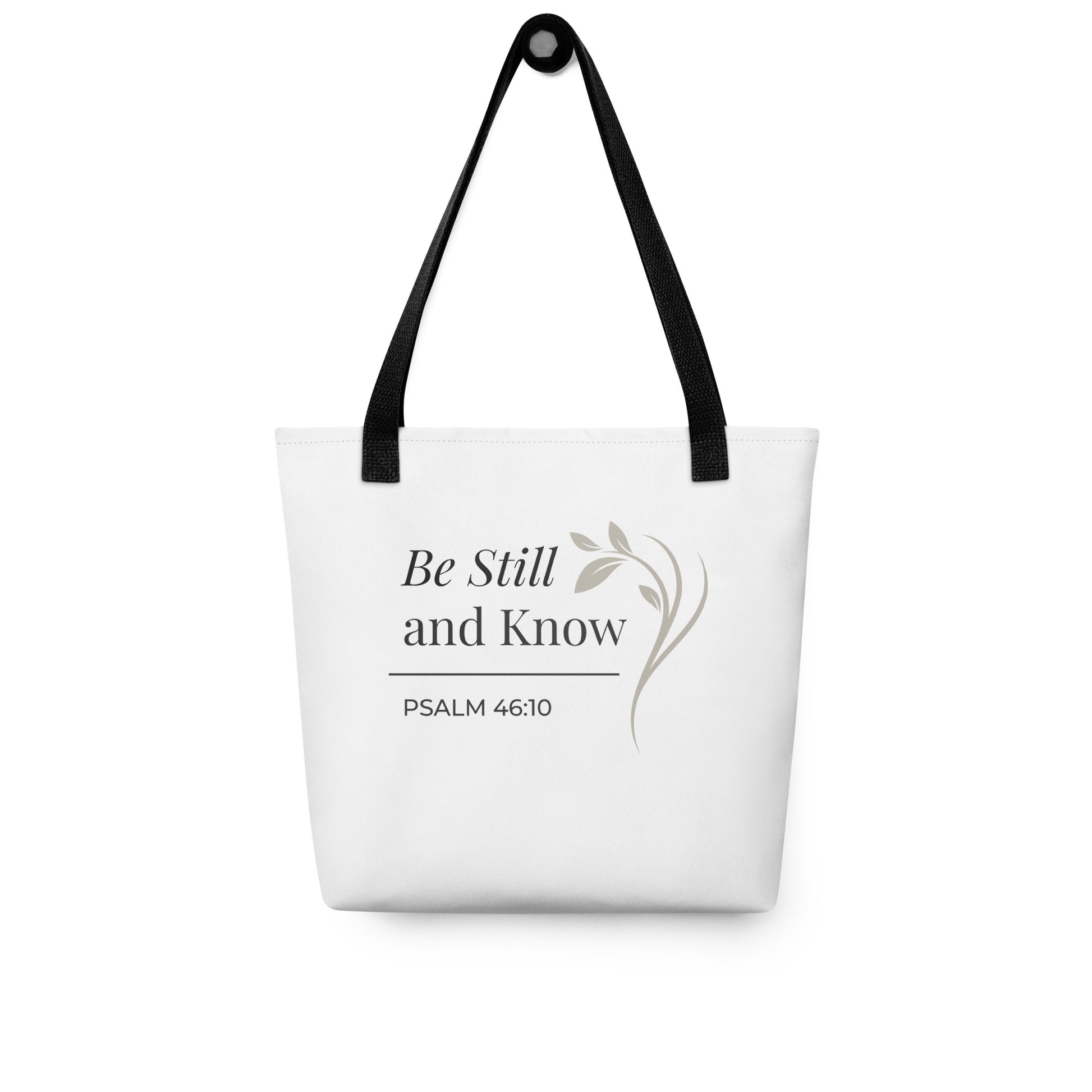 White tote bag hanging on a hook with black handles featuring “Be still and know,” “Psalm 46:10,” and a floral line design in dark gray.