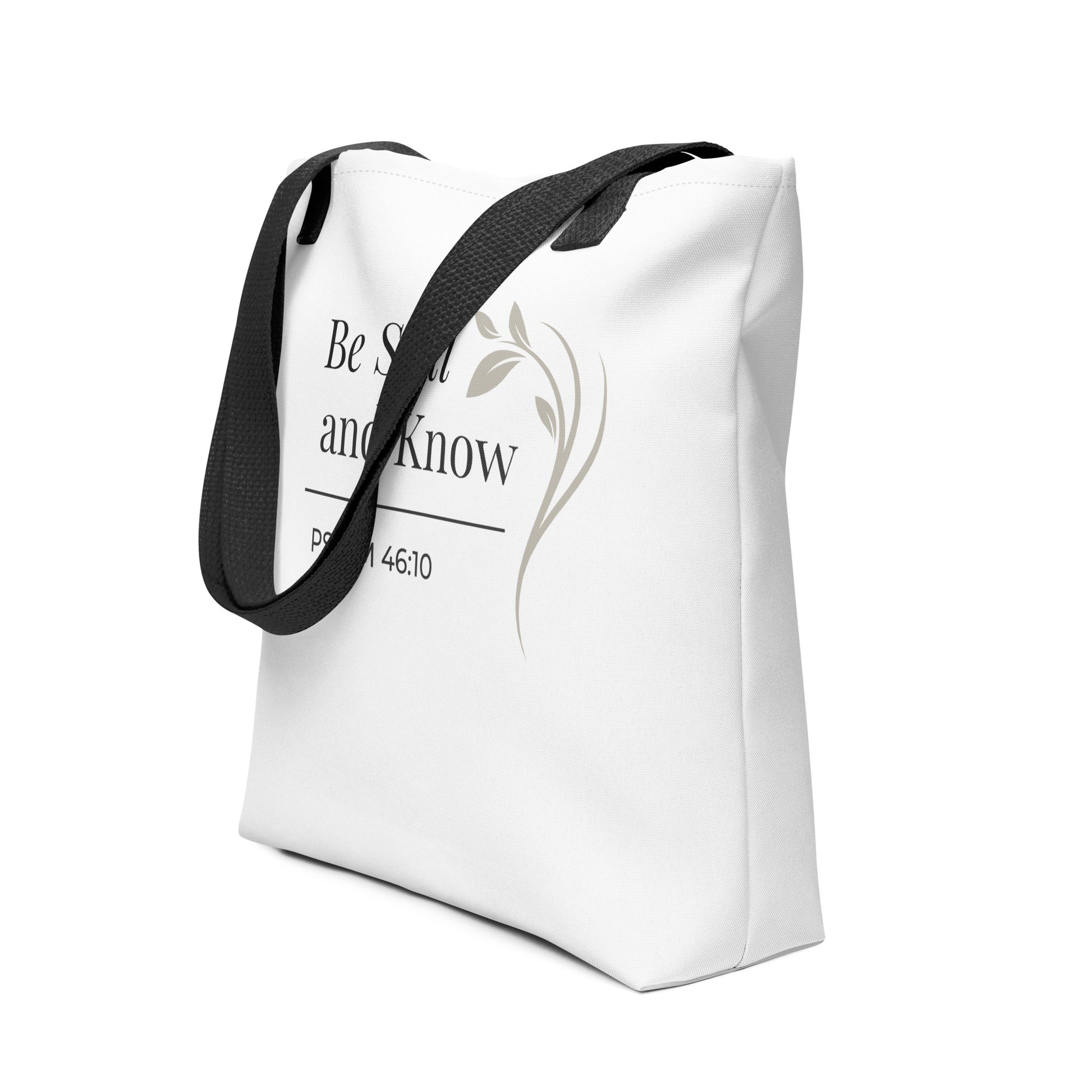 Angled view of a white tote bag with black handles featuring “Be still and know,” “Psalm 46:10,” and a floral line design in dark gray.