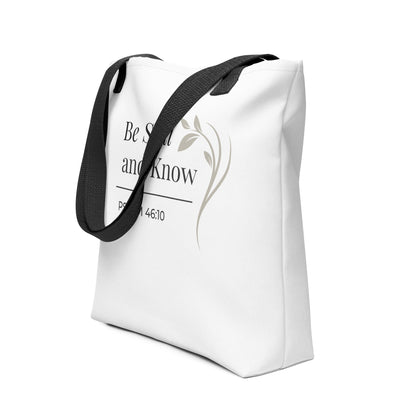 Angled view of a white tote bag with black handles featuring “Be still and know,” “Psalm 46:10,” and a floral line design in dark gray.