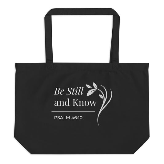 Front view of a large black cotton Christian tote bag with matching black handles and a white “Be Still and Know Psalm 46:10” design.