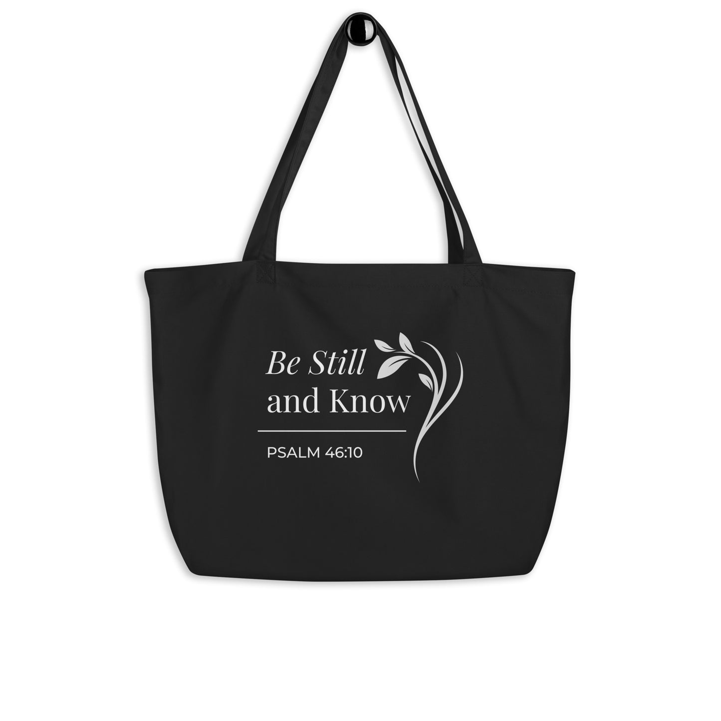 Black Christian large cotton tote bag hanging on a hook, featuring black straps and a minimalist “Be Still and Know” faith design.