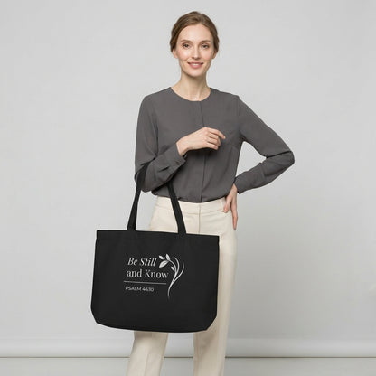 Model carrying a large black cotton Christian tote bag with matching handles and a white “Be Still and Know Psalm 46:10” design.