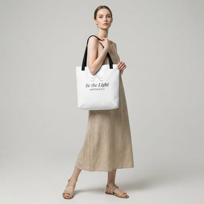 Lifestyle photo of a model holding a white tote bag with black handles featuring “Be the Light,” “Matthew 5:14,” and a sunburst design in dark gray.