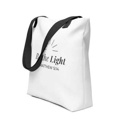 Angled view of a white tote bag with black handles featuring “Be the Light,” “Matthew 5:14,” and a sunburst design in dark gray.