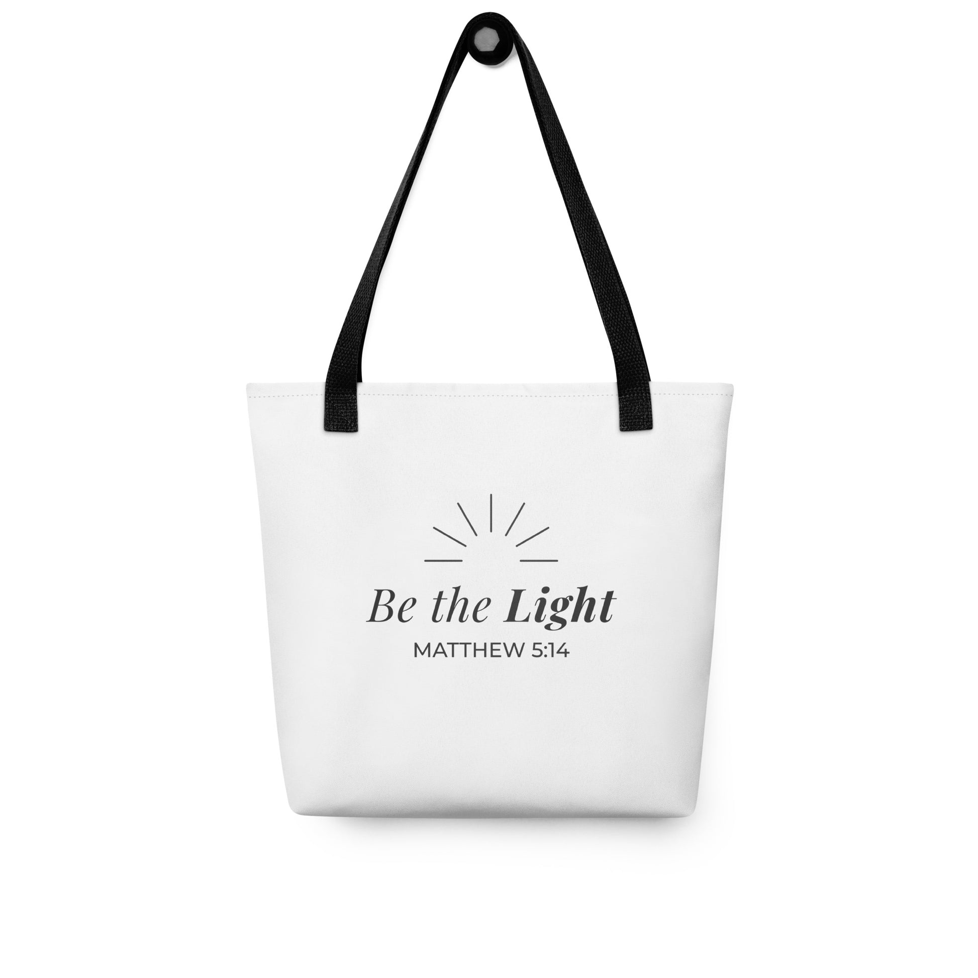 White tote bag hanging on a hook with black handles featuring “Be the Light,” “Matthew 5:14,” and a sunburst design in dark gray.