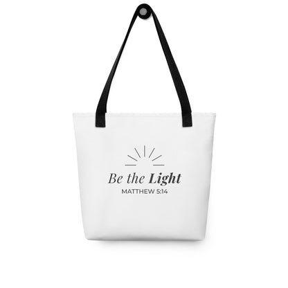 White tote bag hanging on a hook with black handles featuring “Be the Light,” “Matthew 5:14,” and a sunburst design in dark gray.