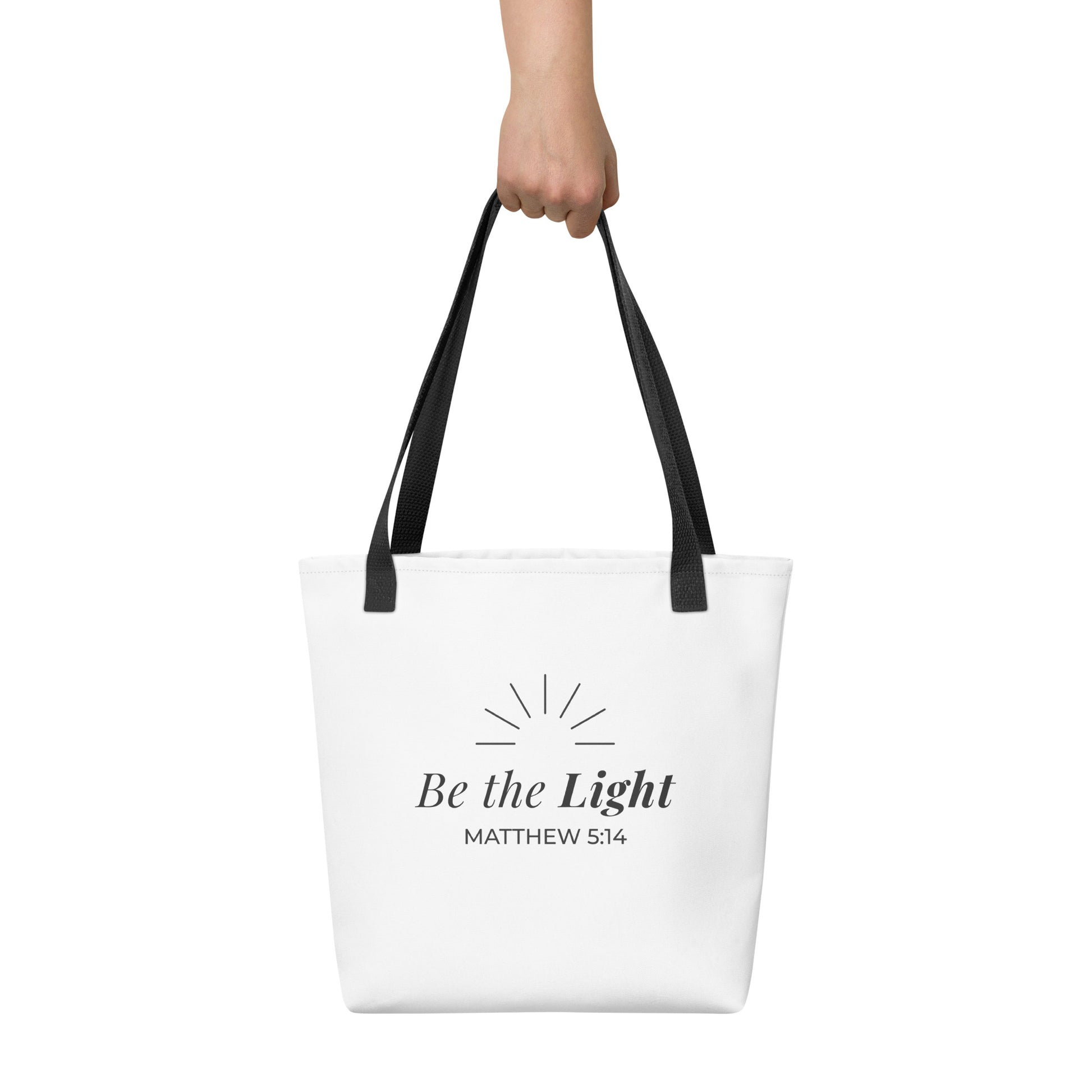 White tote bag held in hand with black handles featuring “Be the Light,” “Matthew 5:14,” and a sunburst design in dark gray.