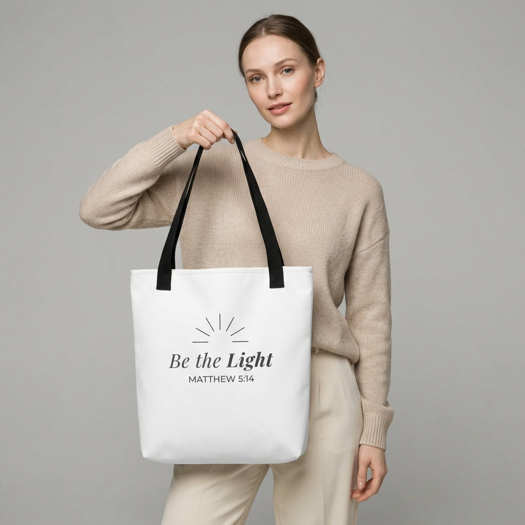 Lifestyle photo of a model holding a white tote bag with black handles featuring “Be the Light,” “Matthew 5:14,” and a sunburst design in dark gray.