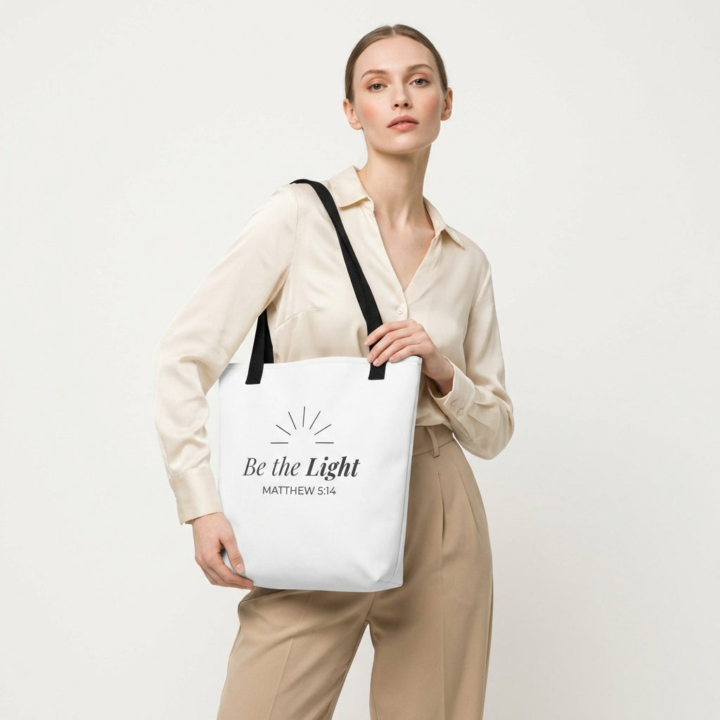 Lifestyle photo of a model holding a white tote bag with black handles featuring “Be the Light,” “Matthew 5:14,” and a sunburst design in dark gray.