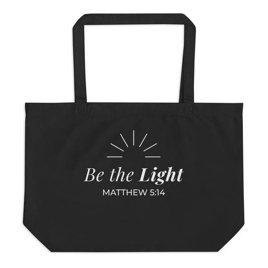 Front view of a black cotton Christian tote bag with matching black handles and a white “Be the Light Matthew 5:14” design.
