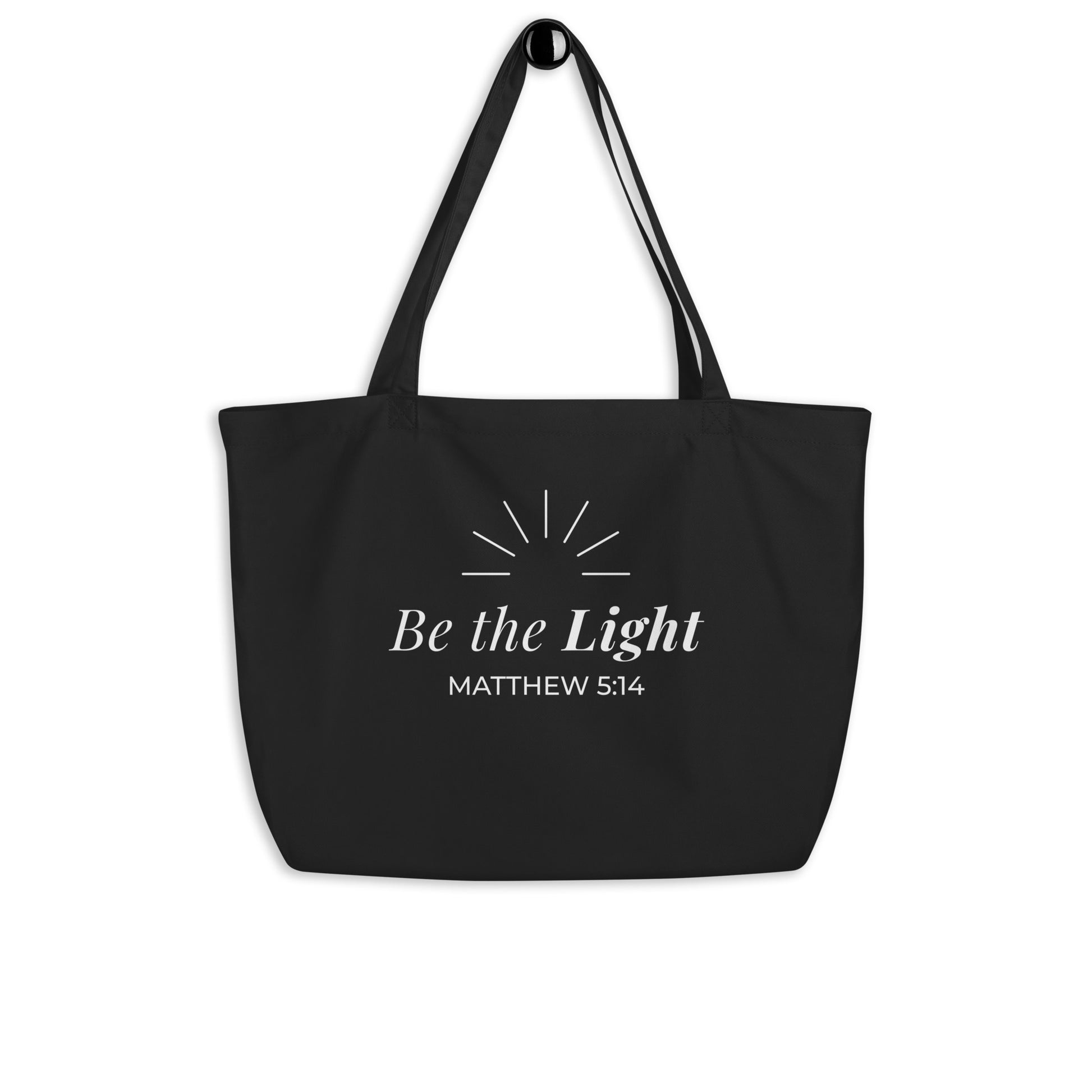 Black Christian cotton tote bag hanging on a hook, featuring matching black straps and a white “Be the Light” faith design.