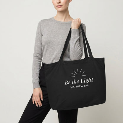 Model carrying a black cotton Christian tote bag with matching black handles and a minimalist “Be the Light Matthew 5:14” design.