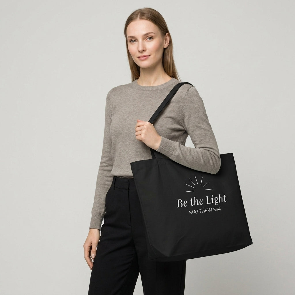 Model carrying a black cotton Christian tote bag with matching black handles and a minimalist “Be the Light Matthew 5:14” design.