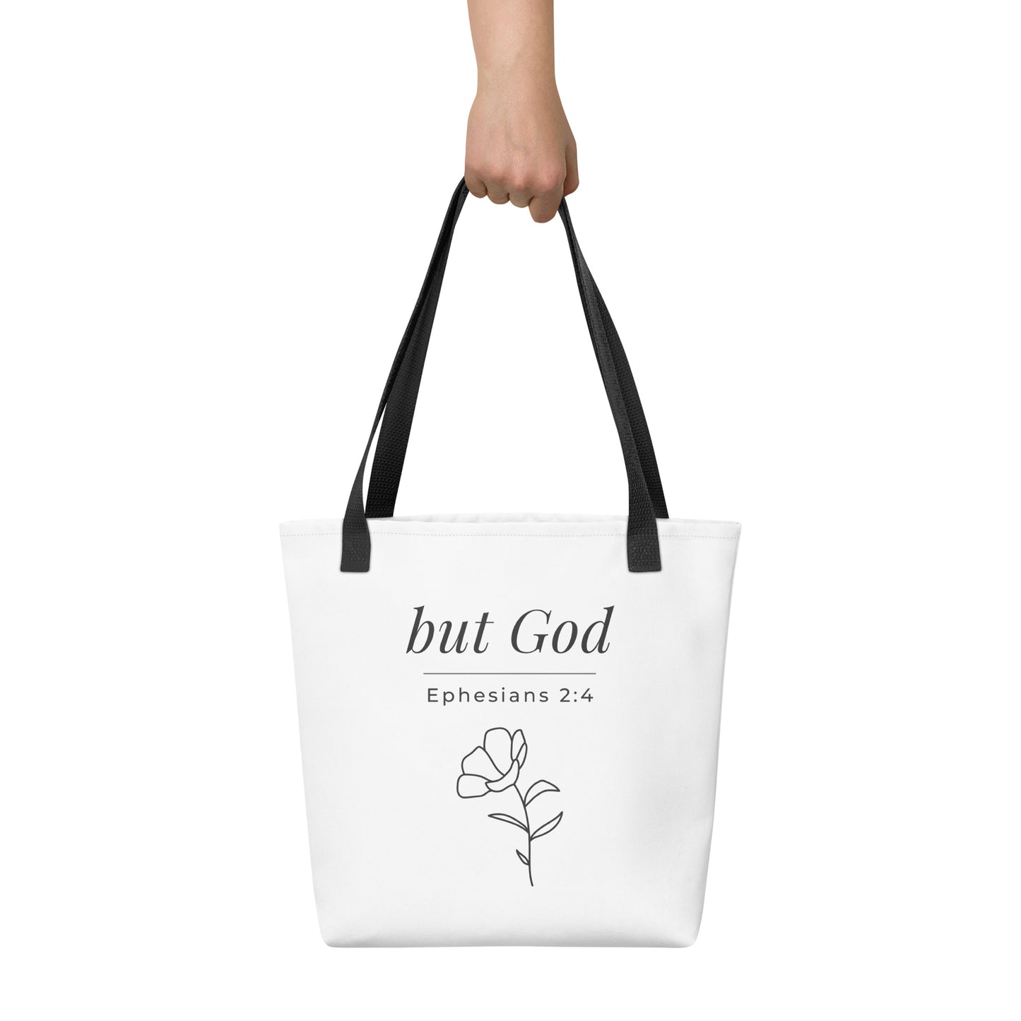 White tote bag held in hand with black handles featuring “but God,” “Ephesians 2:4,” and a flower line-art design in dark gray.
