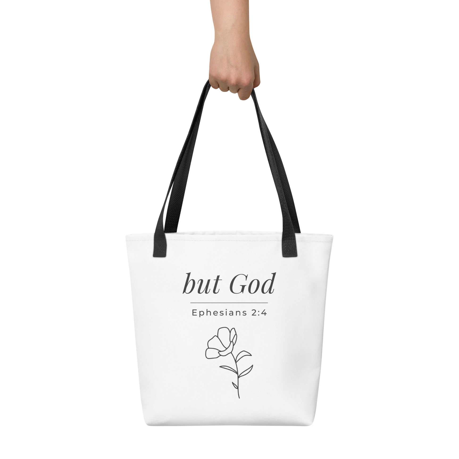 White tote bag held in hand with black handles featuring “but God,” “Ephesians 2:4,” and a flower line-art design in dark gray.