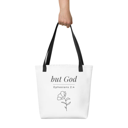 White tote bag held in hand with black handles featuring “but God,” “Ephesians 2:4,” and a flower line-art design in dark gray.