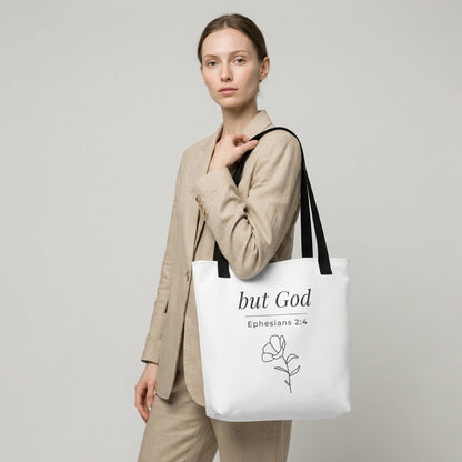 Lifestyle photo of a model holding a white tote bag with black handles featuring “but God,” “Ephesians 2:4,” and a flower line-art design in dark gray.