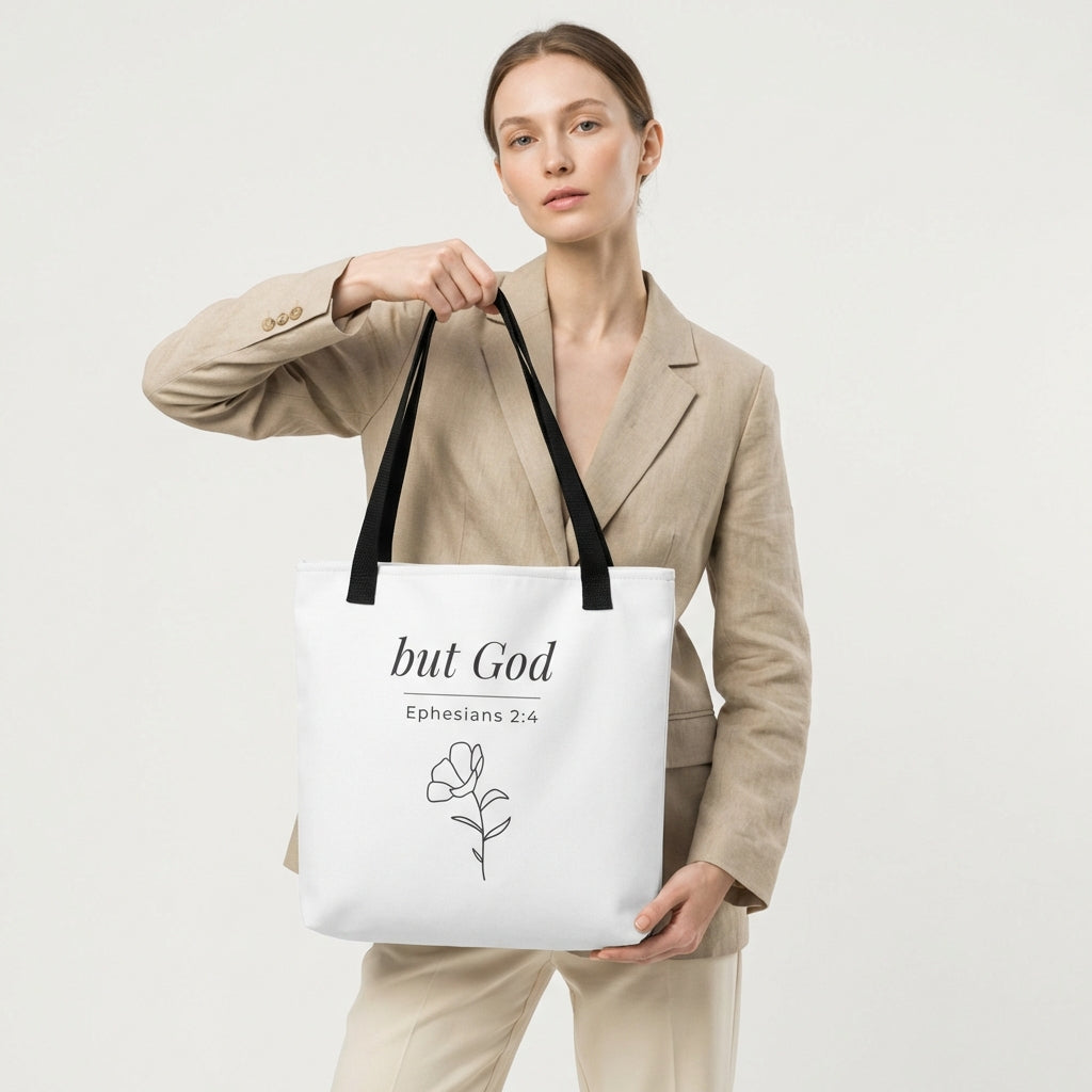 Lifestyle photo of a model holding a white tote bag with black handles featuring “but God,” “Ephesians 2:4,” and a flower line-art design in dark gray.