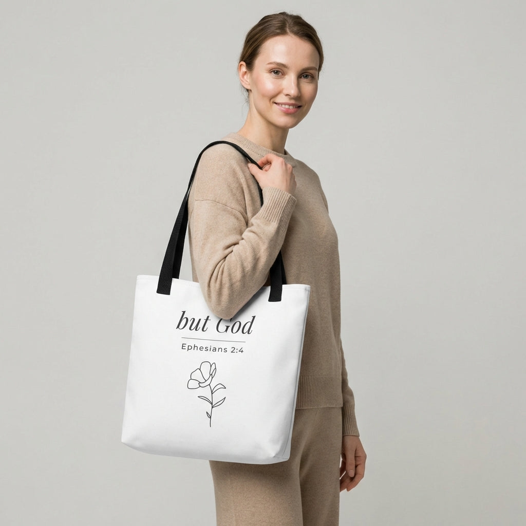 Lifestyle photo of a model holding a white tote bag with black handles featuring “but God,” “Ephesians 2:4,” and a flower line-art design in dark gray.
