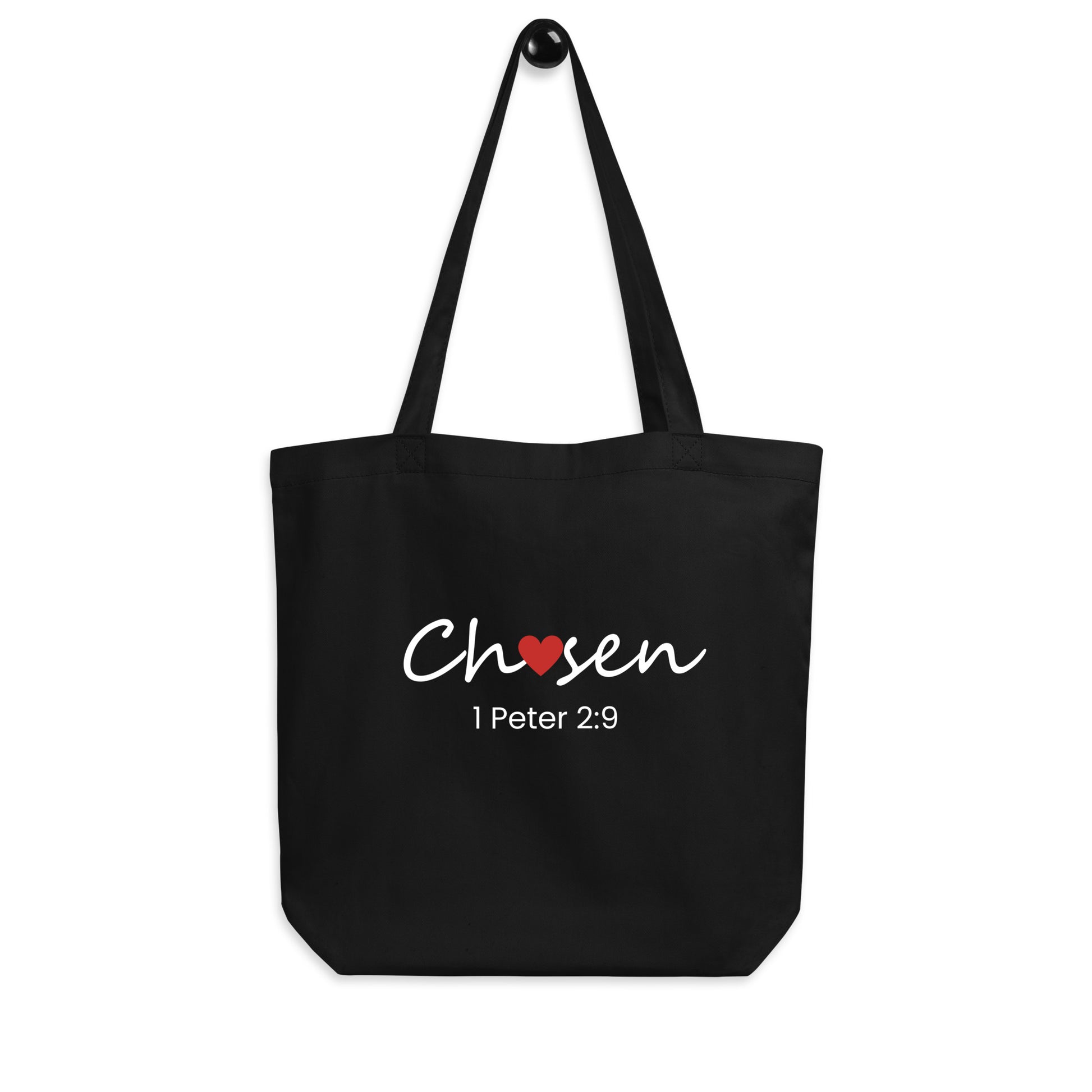 Black cotton tote bag hanging on a hook featuring “Chosen” with a red heart detail and “1 Peter 2:9” below.