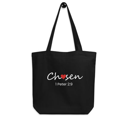 Black cotton tote bag hanging on a hook featuring “Chosen” with a red heart detail and “1 Peter 2:9” below.