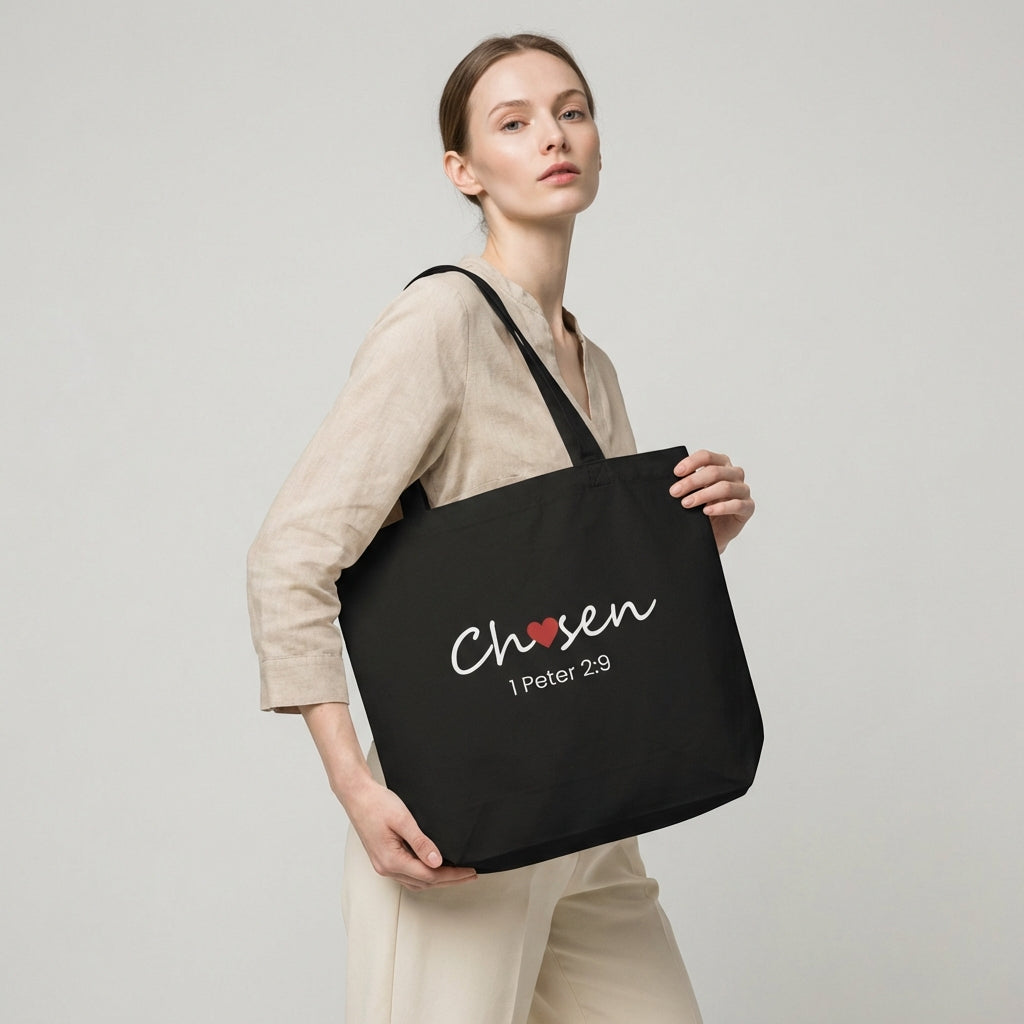 Model holding a black cotton tote bag printed with “Chosen” (red heart accent) and “1 Peter 2:9” beneath.