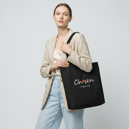 Model holding a black cotton tote bag printed with “Chosen” (red heart accent) and “1 Peter 2:9” beneath.