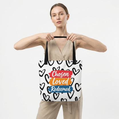Model carrying a white tote with black handles featuring the black heart pattern and “Chosen Loved Redeemed” design.