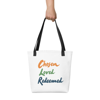 White tote bag held in hand with black straps featuring “Chosen Loved Redeemed” in multicolor script.