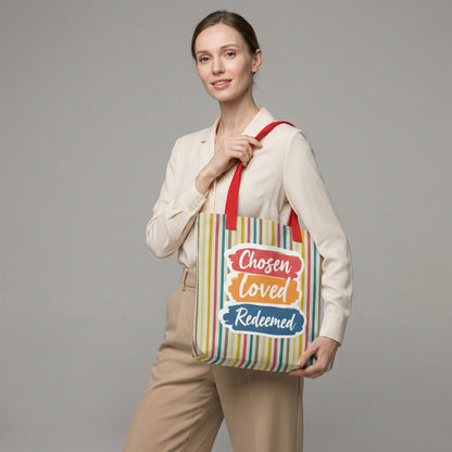 Model holding a striped tote bag with red handles that reads “Chosen Loved Redeemed” in white script on colorful brushstroke blocks.