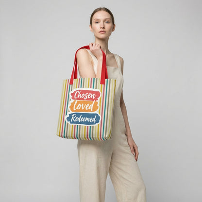 Model holding a striped tote bag with red handles that reads “Chosen Loved Redeemed” in white script on colorful brushstroke blocks.