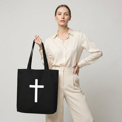 Model holding a black cotton tote bag featuring a large white cross design on the front.