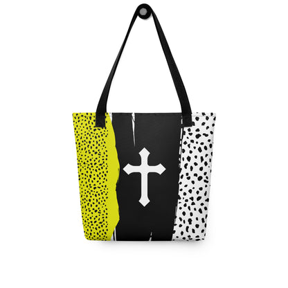 Color-block Christian tote bag with black straps hanging on a hook, displaying a white cross over a black brushstroke background with speckled neon yellow and white panels.