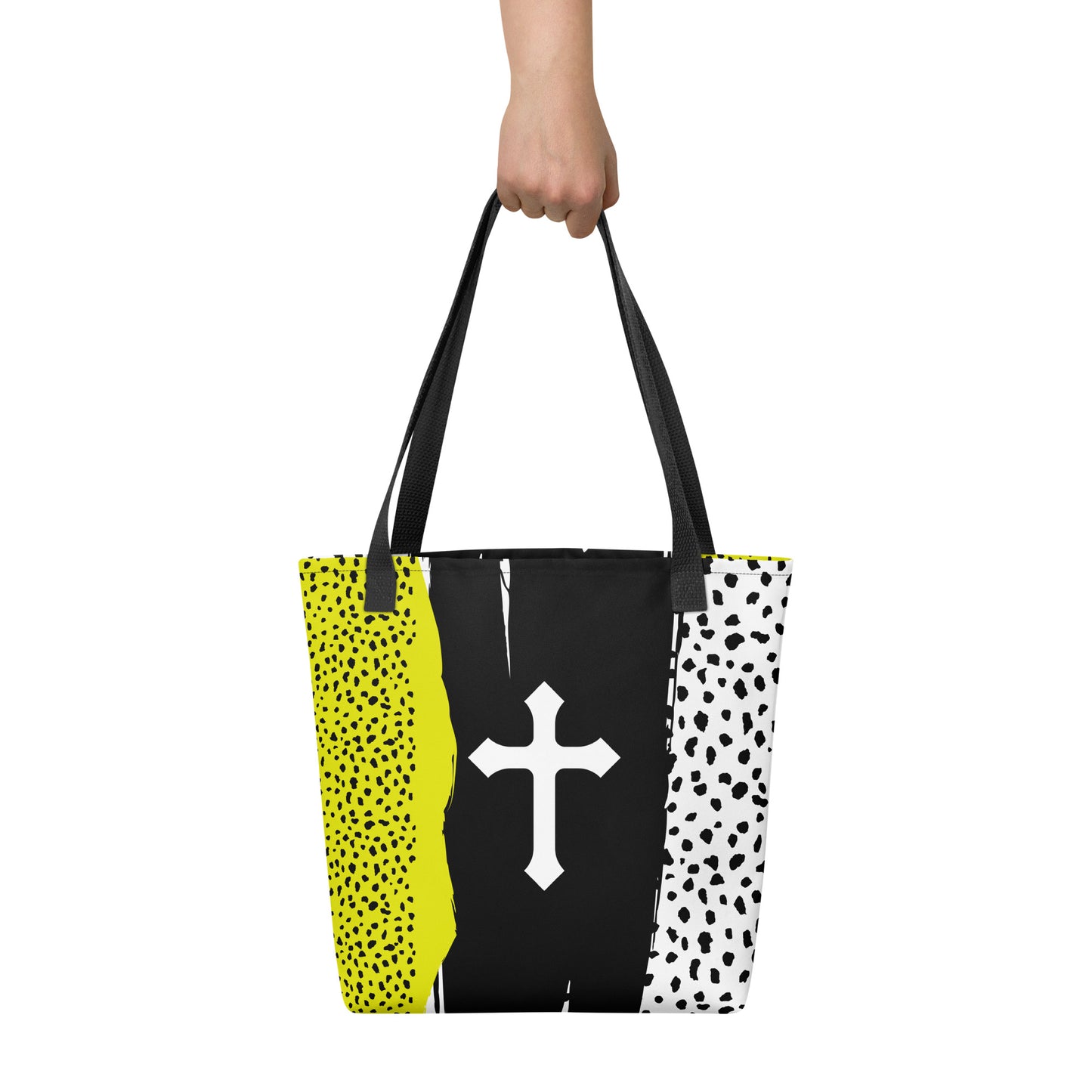 Tote bag held by hand with black straps, featuring a modern color-block pattern with speckled neon yellow and white sides and a white cross on a black center stripe.