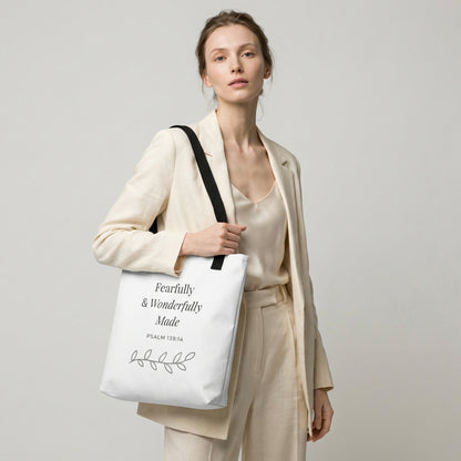 Lifestyle photo of a model holding a white tote bag with black handles featuring “Fearfully & Wonderfully Made,” “Psalm 139:14,” and a leaf vine design in dark gray.