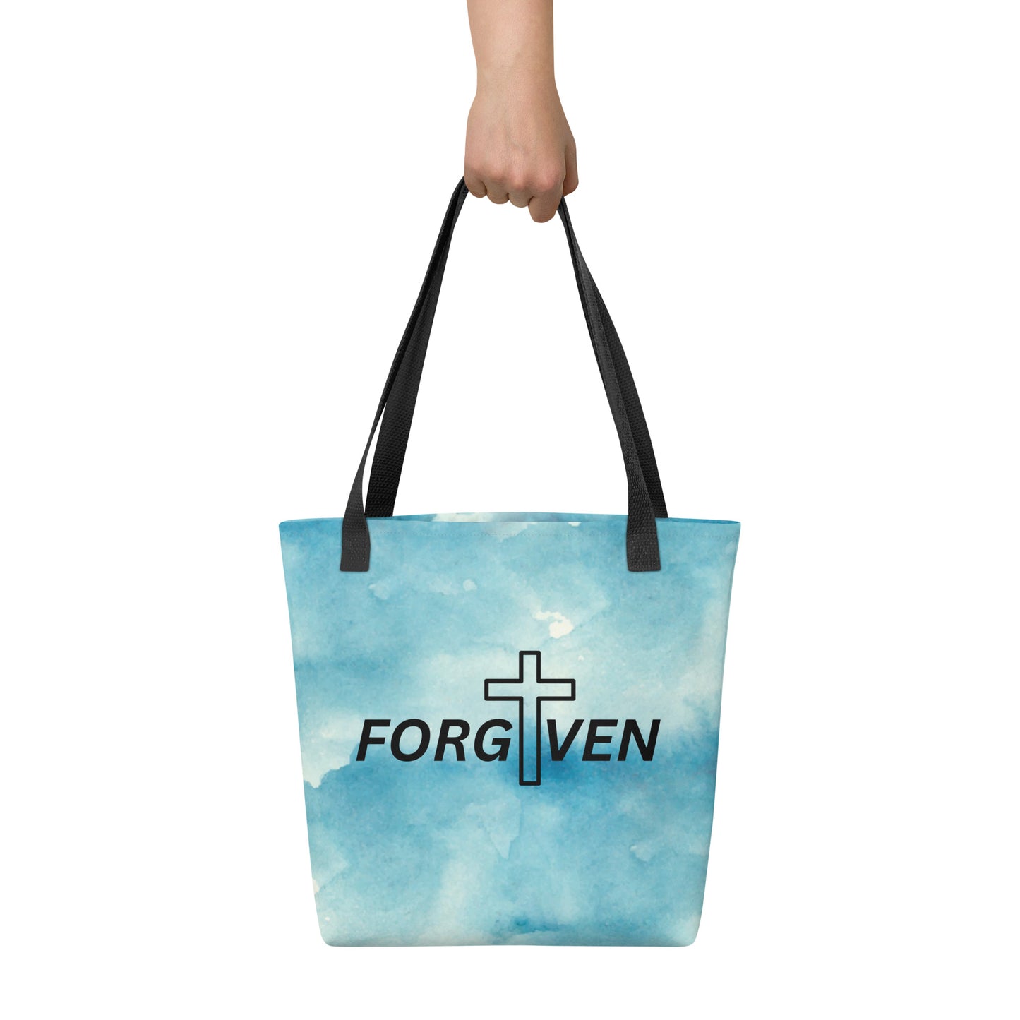 Tote bag carried by hand with black straps, displaying a soft blue watercolor print and bold “FORGIVEN” lettering with a cross.