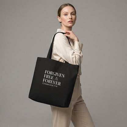 Lifestyle photo of a model holding a black tote bag featuring “Forgiven Free Forever,” “Colossians 1:14,” and a cross design.