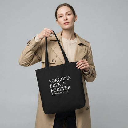 Lifestyle photo of a model holding a black tote bag featuring “Forgiven Free Forever,” “Colossians 1:14,” and a cross design.