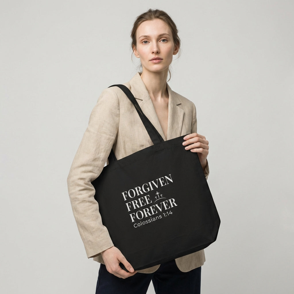 Lifestyle photo of a model holding a black tote bag featuring “Forgiven Free Forever,” “Colossians 1:14,” and a cross design.