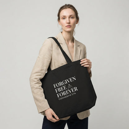 Lifestyle photo of a model holding a black tote bag featuring “Forgiven Free Forever,” “Colossians 1:14,” and a cross design.