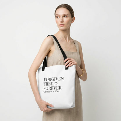 Model holding a white tote bag printed with “Forgiven Free Forever,” “Colossians 1:14,” and a small cross graphic.