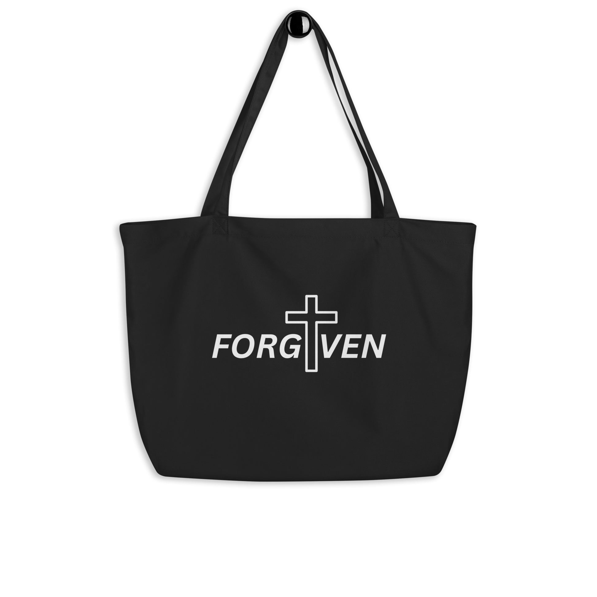 Large black cotton tote bag hanging on a hook featuring “FORGIVEN” with a white cross design on the front.