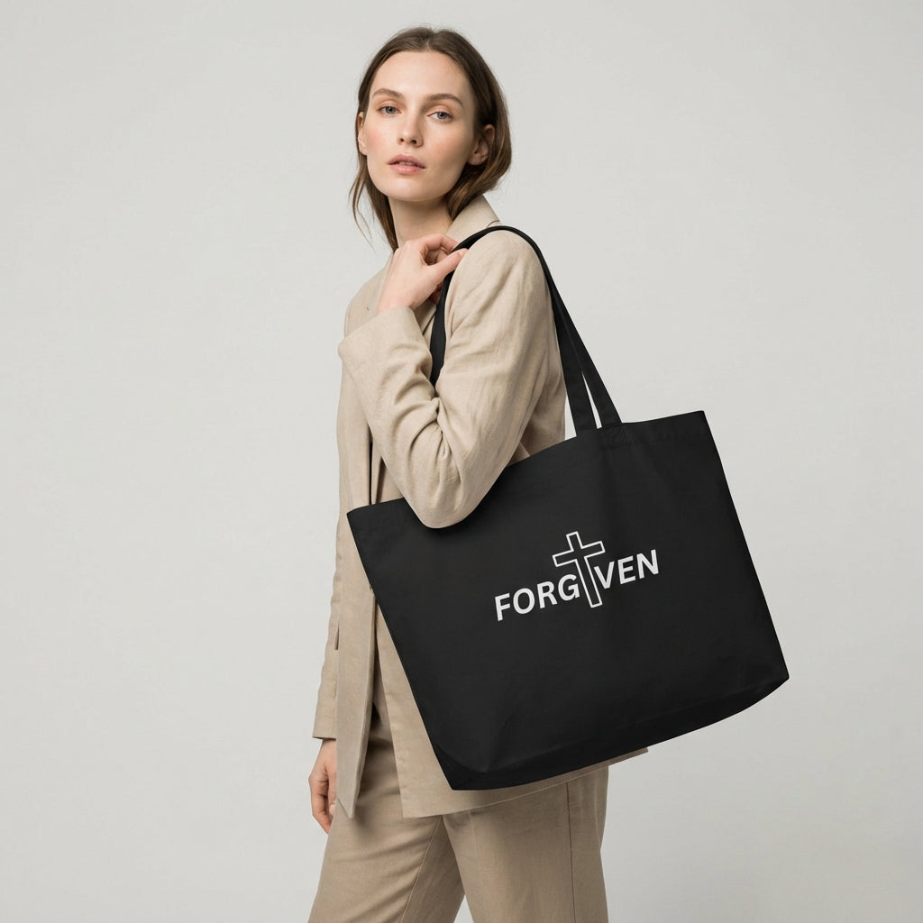 Model holding a large black cotton tote bag with “FORGIVEN” and a white cross graphic on the front.