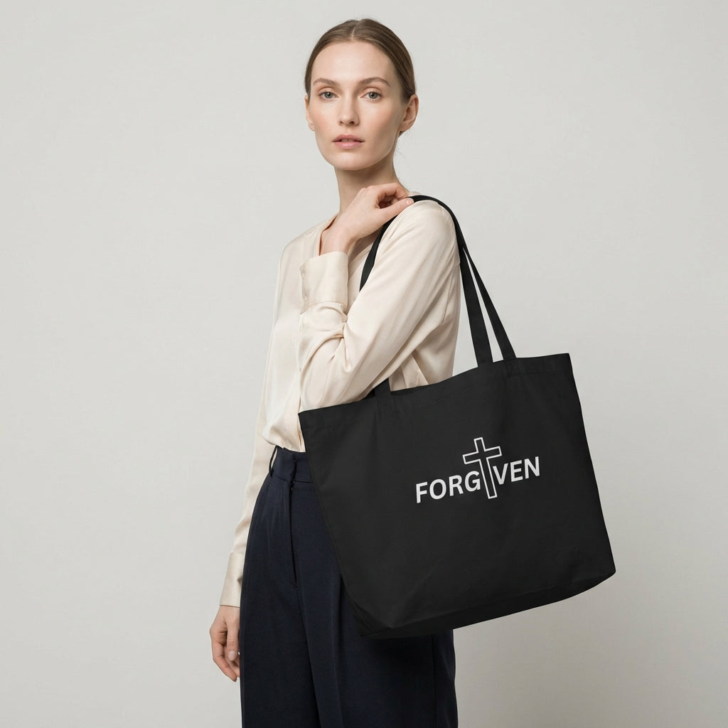 Model holding a large black cotton tote bag with “FORGIVEN” and a white cross graphic on the front.