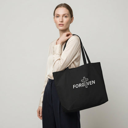 Model holding a large black cotton tote bag with “FORGIVEN” and a white cross graphic on the front.