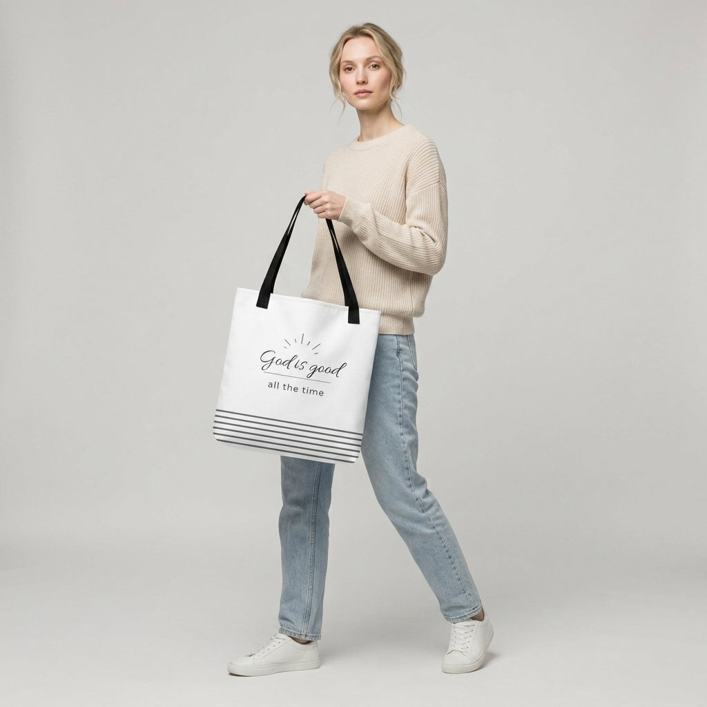 Model holding a white tote bag with black handles that reads “God is good all the time” in dark gray with a striped bottom design.