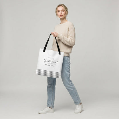 Model holding a white tote bag with black handles that reads “God is good all the time” in dark gray with a striped bottom design.