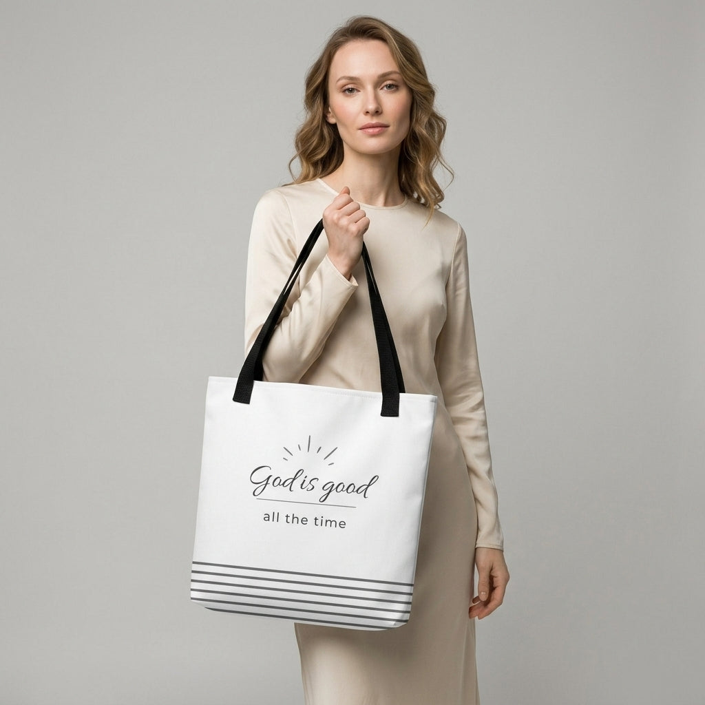Model holding a white tote bag with black handles that reads “God is good all the time” in dark gray with a striped bottom design.